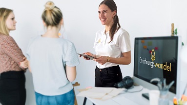 Friedericke Kirsten, Gründerin von Ernährungswandel, steht mit Tablet in der Hand vor zwei Frauen, daneben sichtbar ein großer Monitor mit dem Logo von Ernährungswandel